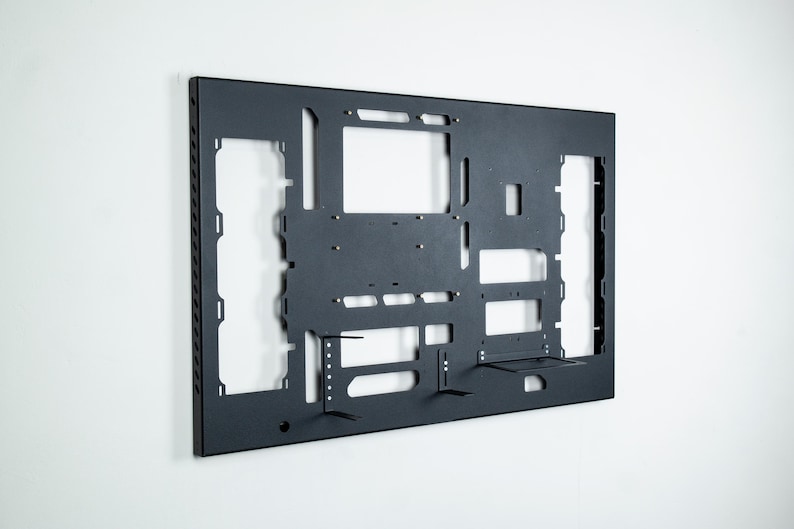 May include: A black metal wall mount with cutouts for mounting computer components. The mount has a textured surface and is designed to be mounted on a wall.
