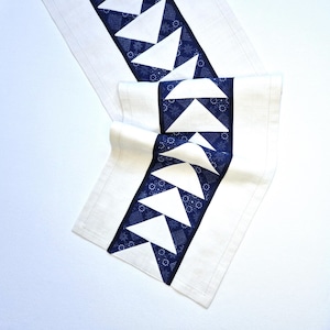 Könnte beinhalten: Weißer Tischläufer mit einem zentralen Design aus blauen und weißen Dreiecken. Die Dreiecke sind in einem sich wiederholenden Muster angeordnet, das von einem dunkelblauen Stoff mit floralem Design umrandet wird. Der Läufer ist aus hellem Stoff gefertigt.