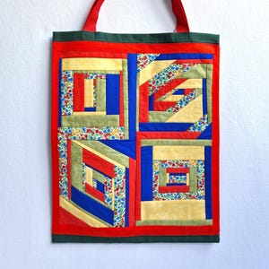 Könnte beinhalten: Eine rechteckige Stofftasche mit einem Patchwork-Design aus vier quadratischen Blöcken. Das Muster besteht aus Streifen in Rot, Blau, Gelb, Olivgrün und einem Blumenmuster. Die Tasche hat rote Stoffgriffe und eine dunkelgrüne Randverzierung.