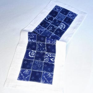 Könnte beinhalten: Ein weiß-blauer Tischläufer. Der Läufer hat ein zentrales Feld aus indigoblauem Stoff, das mit weißen Blumen- und Paisley-Motiven versehen ist und zu einem Patchwork-Design zusammengenäht wurde. Die Ränder sind mit weißem Stoff eingefasst.