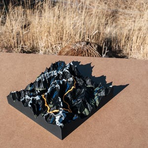 May include: A 3D topographical map sculpture of a mountain range. The black and blue mountains have a yellow trail. The sculpture sits on a brown surface, with a natural background of dry grass and a tree stump.