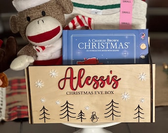 Op de afbeelding: Een kerstavondbox met de naam "Alessis" in rode letters. De box bevat een sokkenaap met een kerstmuts, een boek getiteld "A Charlie Brown Christmas" en een kleine pyjamaset. De box staat op een witte sokkel.
