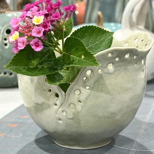 May include: A handmade, light green ceramic vase with a unique, perforated rim. The vase holds a vibrant arrangement of pink and yellow flowers with green leaves. The vase has a smooth, glazed finish and a rounded shape, perfect for displaying small floral arrangements.