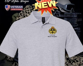 Può includere: Una polo grigio chiaro con maniche corte e colletto. La maglietta presenta uno stemma con la scritta "1-5 CAV Black Knights" e una grafica a stella "NEW". La maglietta ha anche il testo "OP Alpha Designs" e un logo "Veteran Owned Business".