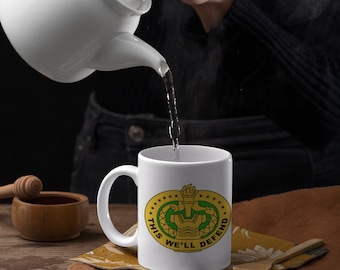 Può includere: Una tazza di ceramica bianca con manico, con un emblema oro e verde con la scritta "THIS WE'LL DEFEND". Una teiera bianca versa acqua nella tazza. La tazza è appoggiata su un tovagliolo giallo fantasia su un tavolo di legno.
