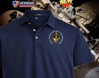 Può includere: Una polo blu navy con un logo ricamato bianco del John F. Kennedy Special Warfare Center and School dell'esercito americano. Il logo presenta uno scudo con una torcia e le parole "US Army JFK Special Warfare Center & School USAJFKSWCg".
