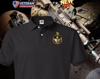Può includere: Polo nero con una toppa ricamata in oro che presenta uno scudo con una torcia e le parole "VIRTUS ET SCIENTIA". La camicia è mostrata su uno sfondo a tema militare.
