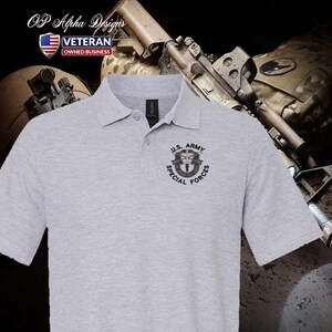 Pu&ograve; includere: Una polo grigia con una toppa ricamata nera che dice "U.S. ARMY SPECIAL FORCES" e presenta un'immagine stilizzata di un soldato con un fucile e un paracadute. La polo &egrave; su uno sfondo mimetico.