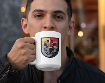 Può includere: Tazza in ceramica bianca con un emblema militare. L'emblema presenta il testo "DE OPPRESSO LIBER" in nero, rosso, giallo e argento. La tazza è tenuta da una persona.