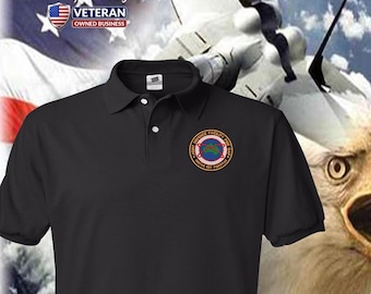 Può includere: Polo nero con una toppa ricamata. La toppa recita "JOINT DEFENCE FACILITY PINE GAP" in un design circolare. Lo sfondo presenta una bandiera americana, un jet da combattimento e un'aquila.