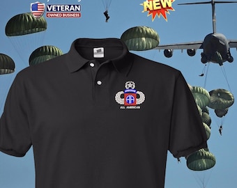 Può includere: Polo nero con una toppa ricamata della 101a Divisione aviotrasportata che dice "Airborne All American". La maglietta è davanti a un cielo blu con paracadute e un aereo militare.