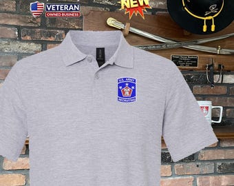 Può includere: Una polo grigia chiara con una piccola toppa di reclutamento dell'esercito americano sul petto. La camicia ha un colletto e una chiusura a tre bottoni. L'immagine include anche il testo "OP Alpha Designs" e "Veteran Owned Business".