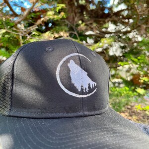 May include: A black and white embroidered wolf howling at the moon on a black mesh trucker hat.