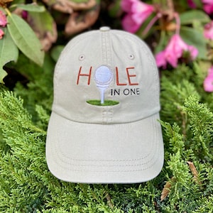 May include: A beige baseball cap with an embroidered golf ball and the text "HOLE IN ONE" in orange.