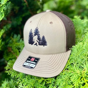May include: A brown and tan trucker hat with a black embroidered image of Bigfoot walking through a forest. The hat has a mesh back and the text "Richardson 112 Trucker Adjustable" on the back.