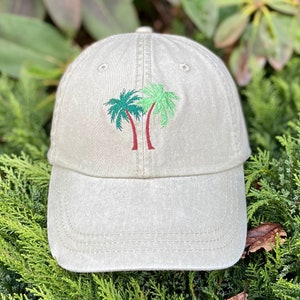 May include: A beige baseball cap with an embroidered design of two green palm trees with red trunks.
