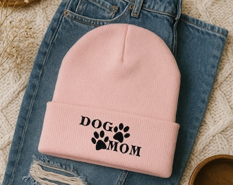 May include: A light pink beanie with the words "DOG MOM" printed in black, along with paw prints. The beanie is resting on a pair of blue jeans.