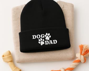 May include: Black beanie with the text "DOG DAD" in white, featuring paw prints. The hat rests on a tan sweater, with a dog bone and a rope toy with orange and white accents.