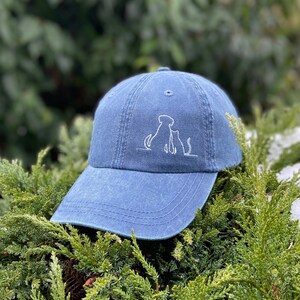 May include: A blue denim baseball cap with white embroidery of a dog and cat sitting together.
