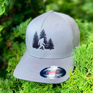 May include: Gray baseball cap with an embroidered design of a Bigfoot walking through a forest. The cap has a Flexfit logo and the text "The Original Flexfit S/M It Fits Your Life".