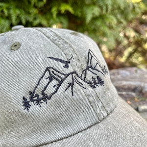 May include: A green baseball cap with an embroidered design of mountains and a bird. The design is in black thread.