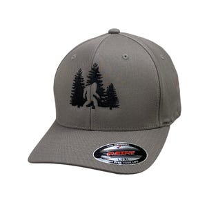 May include: Gray Flexfit baseball cap featuring a black embroidered design of Bigfoot walking in front of two pine trees. The cap has a curved brim and a Flexfit label that says "IT FITS YOUR LIFE."