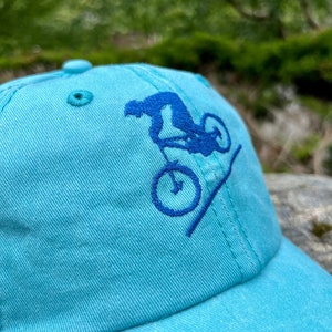 May include: A turquoise blue baseball cap with an embroidered design of a person riding a bicycle on a ramp. The design is in blue thread.