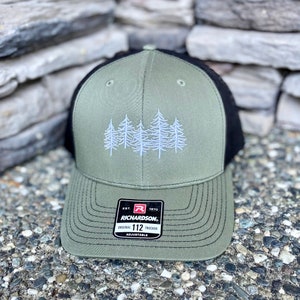 May include: A green and black trucker hat with an embroidered design of five white pine trees. The hat has a black mesh back and a green front with a black brim. The hat has a black adjustable strap with a label that reads "EST. 1970 RICHARDSON. ORIGINAL 112 TRUCKER ADJUSTABLE."