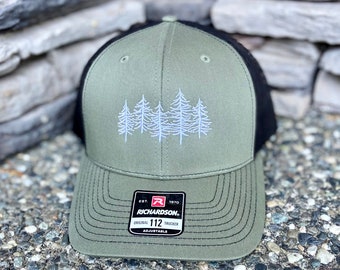 May include: A green and black trucker hat with an embroidered design of five white pine trees. The hat has a black mesh back and a green front with a black brim. The hat has a black adjustable strap with a label that reads "EST. 1970 RICHARDSON. ORIGINAL 112 TRUCKER ADJUSTABLE."