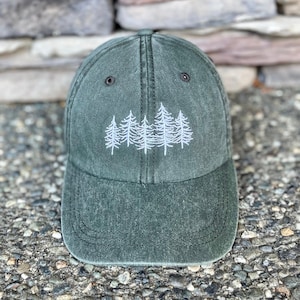 May include: A dark green baseball cap with a white embroidered design of pine trees. The cap has a curved brim and a classic six-panel design. The hat is made of a soft, washed cotton material.