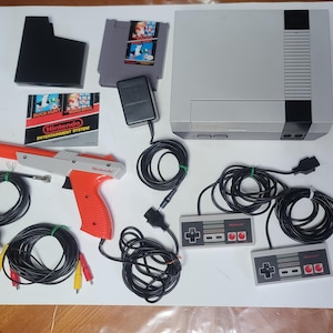 Puede incluir: Un conjunto completo de Nintendo Entertainment System (NES), que incluye la consola, dos mandos, una pistola Zapper, cartuchos de juego y cables de conexión. La consola es gris con detalles en negro. La pistola es naranja y gris.