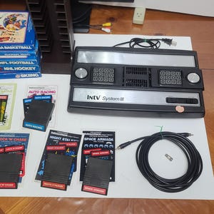 May include: A vintage black and silver INTV System II video game console with several game cartridges, including NBA Basketball, NFL Football, and NHL Hockey. Also included are game cartridges for Auto Racing, Lock 'N Chase, and Space Armada.