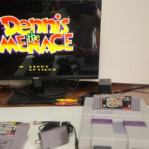 May include: A Super Nintendo Entertainment System (SNES) console with two game cartridges and a monitor displaying the game "Dennis the Menace." The console is gray with purple accents. The game cartridges are gray.