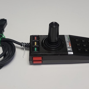 May include: A black joystick with a red button and a numeric keypad. The joystick has a black and silver base with the text "CASSETTE  FUN  II" printed on it. The joystick is connected to a black cable with a connector on the end.