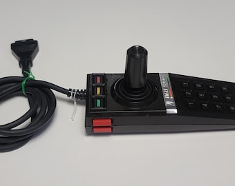 Puede incluir: Un joystick negro con un botón rojo y un teclado numérico. El joystick tiene una base negra y plateada con el texto "CASSETTE  FUN  II" impreso en él. El joystick está conectado a un cable negro con un conector en el extremo.