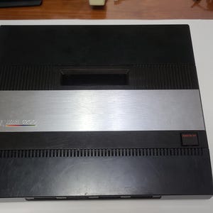 May include: Black Atari 5200 video game console. The console has a silver stripe across the middle with the Atari logo and model number. The top and bottom are black with ventilation slots. A red power button is on the right.