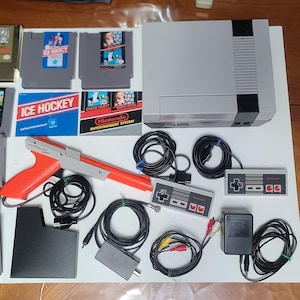 May include: A complete Nintendo Entertainment System (NES) set, including the console, controllers, game cartridges, and accessories. The set includes a grey console, a red and grey light gun, and various game cartridges with colourful labels. The set is displayed on a white surface.