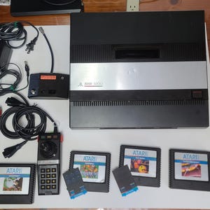 May include: An Atari 2600 video game console with controllers, power supply, and game cartridges. The console is black and silver, with the Atari logo. The cartridges feature colorful game labels. The controllers are black with a joystick and keypad.