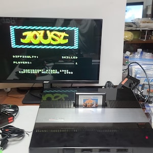 May include: An Atari video game console with a game cartridge inserted. The screen displays the game "Joust". Two black joysticks are in the foreground. The console is on a table with game cartridges and game packaging.