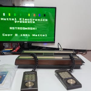 May include: A vintage video game console with two controllers and a monitor displaying "ASTROSMASH!" from Mattel Electronics. The console has a brown and black design, and the controllers are black with silver accents. A game cartridge is visible.