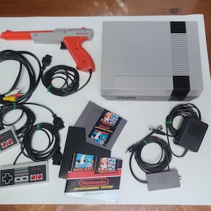 May include: A grey and black Nintendo Entertainment System console with two rectangular controllers, a grey and orange light gun, a Duck Hunt game cartridge, a power adapter, AV cables, and a Nintendo Entertainment System instruction manual.