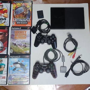 Puede incluir: Una consola PlayStation 2 con dos mandos negros, discos de juegos y varios cables. Los discos de juegos incluyen títulos como "NCAA Football 2004", "Deer Hunt 2005 Season", "NASCAR Thunder 2003", "Secret Weapons Over Normandy", "Madden 06" y "SeaWorld Deep Sea Adventure".