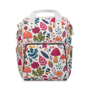 May include: A backpack featuring a white background with a repeating pattern of colorful mushrooms, pink and orange flowers, green leaves, and brown acorns. The bag includes a top handle, a front zippered pocket, and side pockets with tan trim.