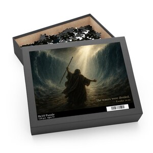 May include: A black puzzle box featuring a biblical scene of a figure with outstretched arms, standing between walls of water. The box reads "The waters were divided. Exodus 14:21" and "8x10 Puzzle 120 pcs Ages 9+". The puzzle measures 20 x 25 cm.