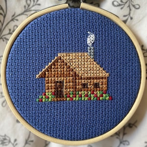 May include: A cross-stitch embroidery of a brown cabin with a chimney and a flower bed, set against a blue background. The embroidery is framed in a wooden hoop. The cabin has a detailed roof and door.