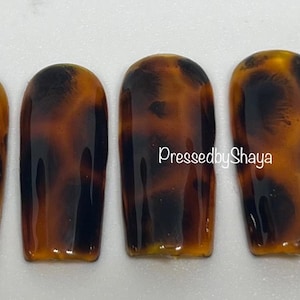 May include: Set of six tortoise shell press-on nails. The nails are a medium length and have a rounded shape. The nails are a brown and black color with a glossy finish.  PressedbyShaya.