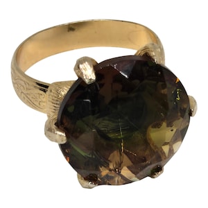 May include: A gold ring featuring a large, round, faceted gemstone with shifting hues of brown, green, and yellow. The band is crafted with an engraved floral pattern and the stone is secured by six prongs that resemble small, textured claws.