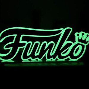 May include: A glowing green and black Funko logo, with the brand name in a stylized font and a crown icon. The neon-like sign is set against a dark background, creating a striking visual effect.