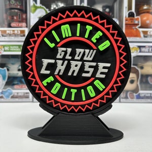 May include: A black, circular display piece with the words "LIMITED GLOW CHASE EDITION" in green and silver lettering. The text is set against a black background with a red zig-zag border. The display is mounted on a black stand.