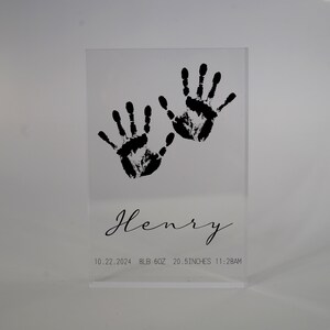 Op de afbeelding: Een heldere acrylblok met zwarte handafdrukken en de naam "Henry" gegraveerd. De datum "10.22.2024", het gewicht "8 LB 6OZ", de lengte "20.5 INCHES" en de tijd "11:28AM" zijn ook op de blok gedrukt.